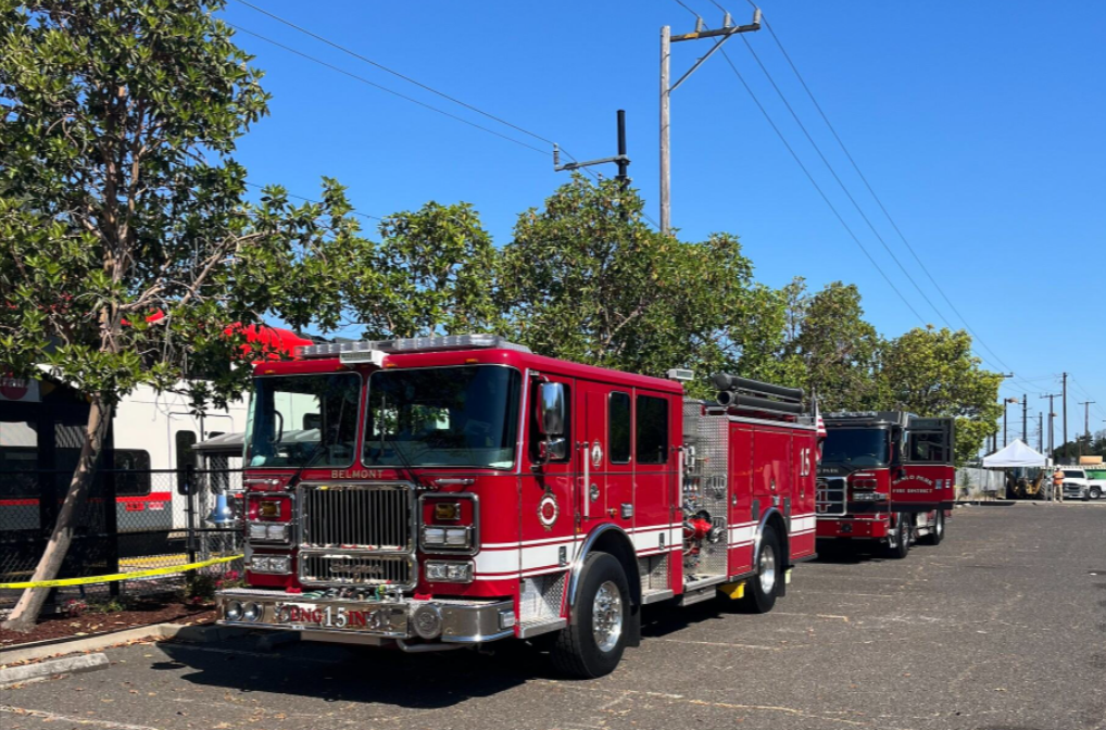 Two dark red firetrucks are parked in a parking lot, parallel to Caltrain's new electric train halted on the tracks.