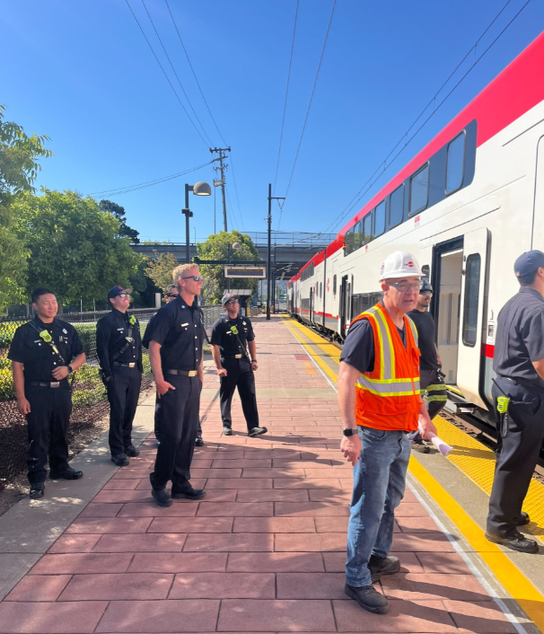 A group of firefighters observe Caltrain's new electric train at the Hayward Park Station, learning about the new safety features on a bright sunny day.