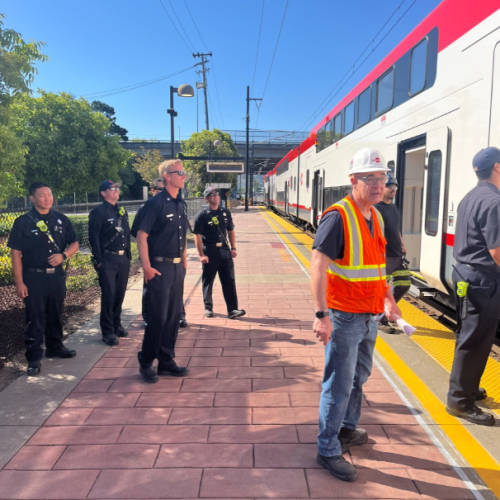 A group of firefighters observe Caltrain's new electric train at the Hayward Park Station, learning about the new safety features on a bright sunny day.